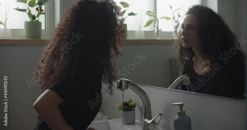 An insecure woman staring at herself while water running in the sink. Slow motion, handheld, medium shot.  photo