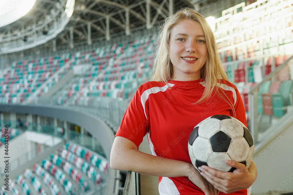 Portrait of young caucasian woman in red football strip holding ball at big soccer stadium smiling to camera Stock Foto Adobe Stock