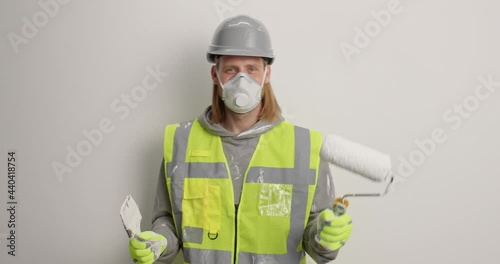 Positive redhead man dances with painting tools going to begin house reapir moves against white background wears protective helmet respirator and uniform paints walls in new house. Maintenance photo