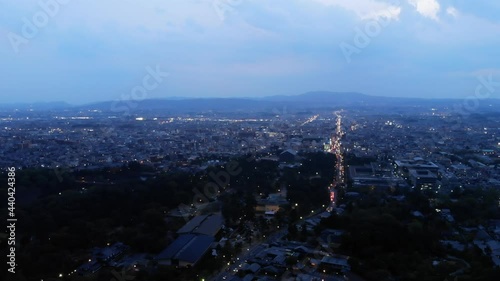 Aerial panorama of Nara city at late evening, bright lights on roads, dim park area seen below, buildings in dusk at distance. Blue and cloudy sky, mountains far away photo