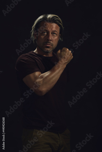Dark portrait of a middle-aged man with blond hair and a gray stubble beard in a dark brown t-shirt showing his muscular arm. © ysbrandcosijn