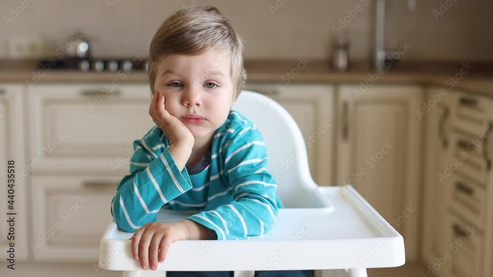 Little hungry and sad boy sitting in white baby chair in kitchen room and waiting some food from parents Little hungry and sad boy sitting in white baby chair in kitchen room and waiting some food from parents