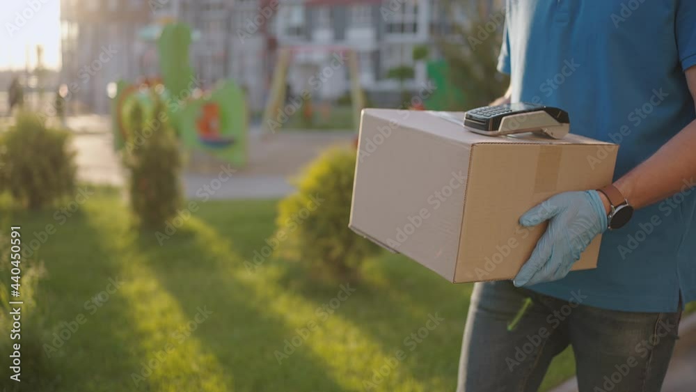A male deliveryman in gloves brings a box and gives the terminal to pay for the purchase. Carry a box with an NFC credit card payment terminal or mobile phone. Contactless payment