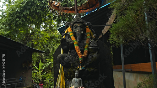 An old stone statue of Ganesha covered with moss with a wreath of yellow flowers on his neck standing on the street © Nicolas Gregor