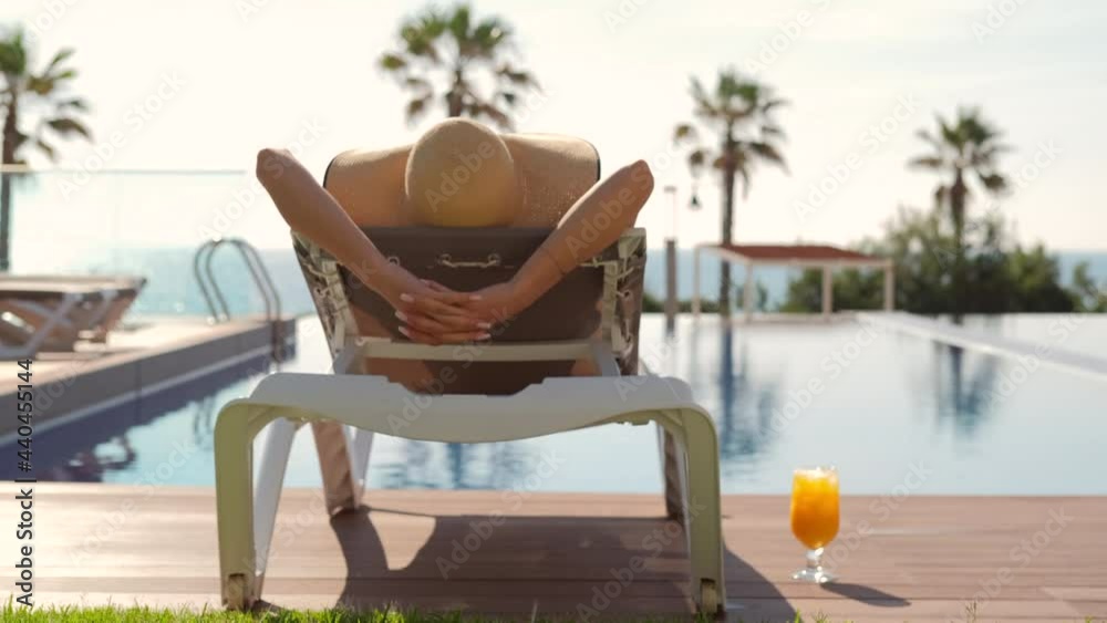 Rear view woman wear straw hat lying on deckchair on poolside put hands behind head relaxing take sun bath, sea, palm tree, empty swimming pool scenery on background. Summer holidays, travel concept