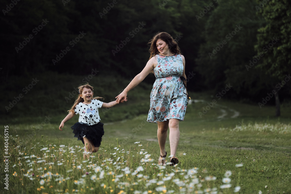 Have fun with child in nature, the joy of motherhood. Young beautiful European mother in blue dress runs through chamomile field with her little daughter and laughs.