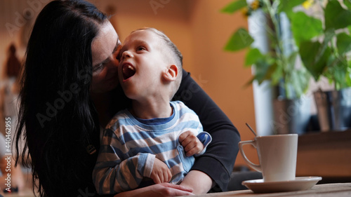 Close up portrait of a little boy with special needs and mom laughing at a table in a cafe, lifestyle. Mom's love for her child, inclusion.Happy disability kid concept