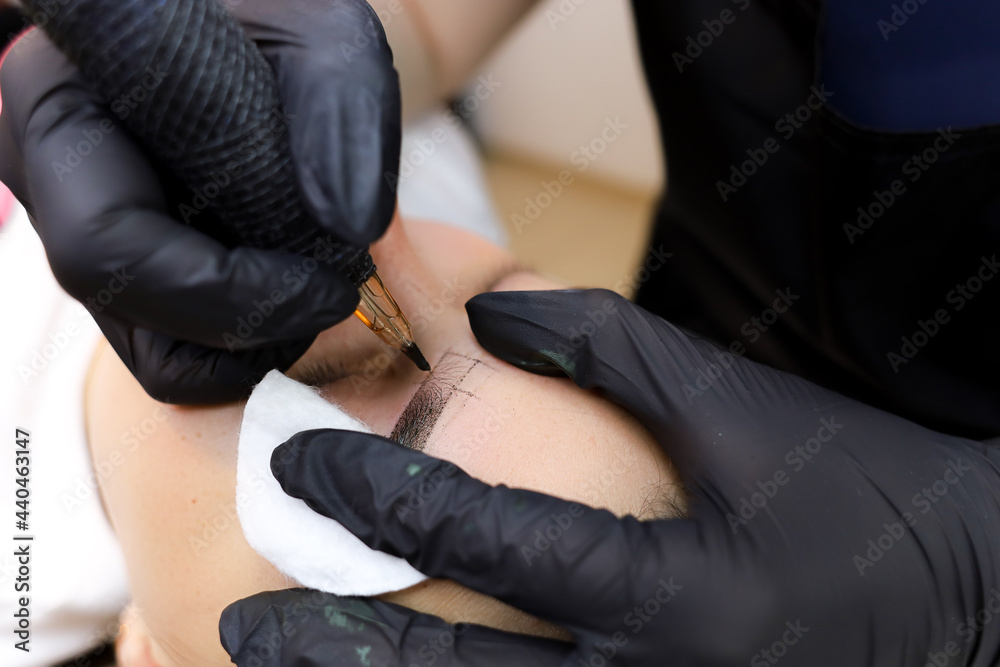 the master of permanent makeup performs a tattoo of the eyebrows with a tattoo machine holding the eye area with a cotton sponge