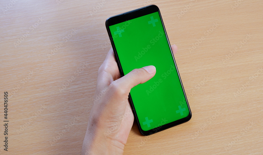 Close-up of a woman's hand holding a phone with a green screen on the background of a wooden table. chromakey.