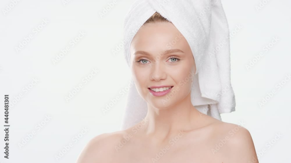 Gorgeous young Caucasian slim woman with towel on her hair touches her face after bathroom turning to the camera and smiling wide for it on white background | Face cream advertising