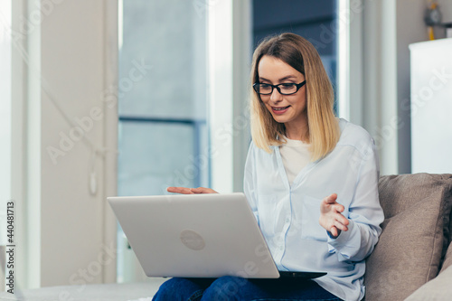 Beautiful blonde woman in casual clothes sitting on a sofa at home, and uses a laptop for video communication with friends and family