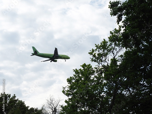 Airliner descends by releasing landing gear over green trees against the backdrop of a cloudy sky
