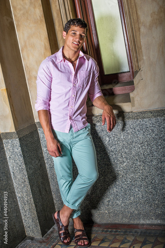 Dressing in a light pink shirt, light green pants and dark brown sandals, a young handsome guy is standing by a window and smiling..
