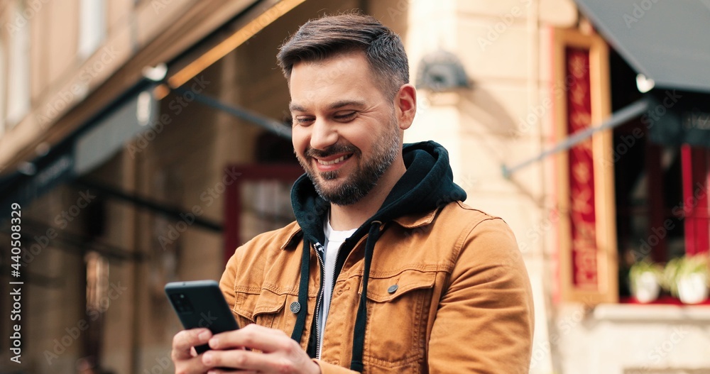 Portrait of caucasian man with dark hair out and about in the city streets during the day, looking at smartphone and smiling while chatting with his friends. Portrait of caucasian man with dark hair out and about in the city streets during the day, looking at smartphone and smiling while chatting with his friends.