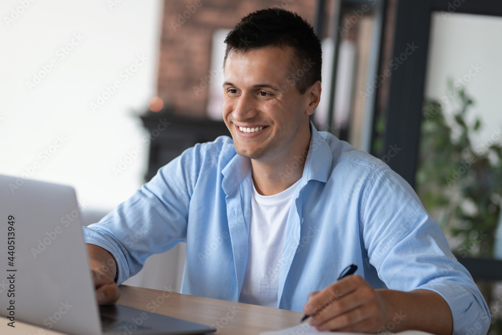 Caucasian man student, using a laptop, studying remotely from home, taking notes on notepad during online lesson, distance education concept, smiling Caucasian man student, using a laptop, studying remotely from home, taking notes on notepad during online lesson, distance education concept, smiling