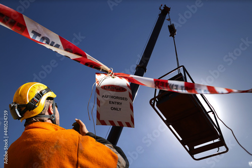 Safety work practice red and white warning danger tag tape sign applying on exclusion dropped zone where rigger operating 2 way radio with crane operator during lifting heavy load at open field 
