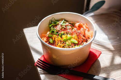 Healthy poke bowl dish with salmon and vegetables in kraft paper packaging on a wooden table. Takeout food. Fitness food. Dinner.