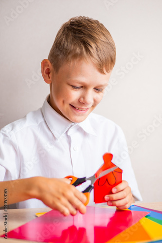 A smiling blond boy in a white cotton shirt is cutting a little man out of red paper with orange scissors