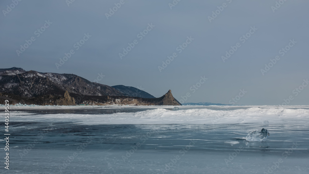 Snowdrifts lie on the smooth surface of the frozen lake. In the foreground is a bizarre shard of ice. Glare on the edges. Picturesque mountain range against the blue sky. Baikal