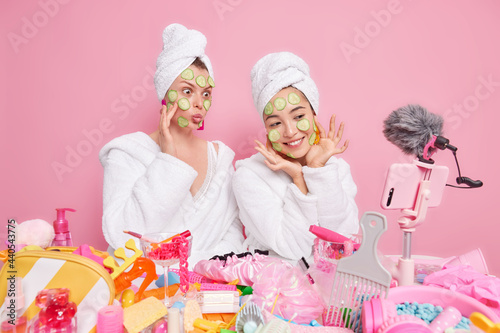 Two women bloggers show how to make natural face mask apply cucumber slices on face record vlog video on smartphone wear white soft bathrobes and towels over head isolated over pink studio wall