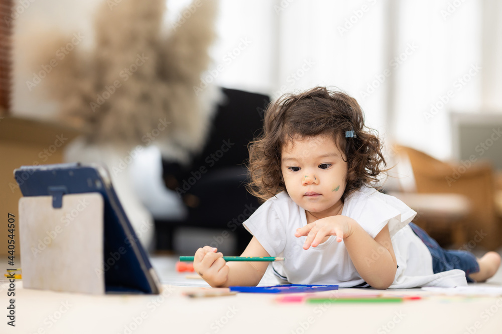 Naklejka premium Cute daughter painting coloring with watercolor on the large paper Learning by tablet. Sitting on the floor in living room. Happy kid moment in the house. Art Activity in kindergarten.