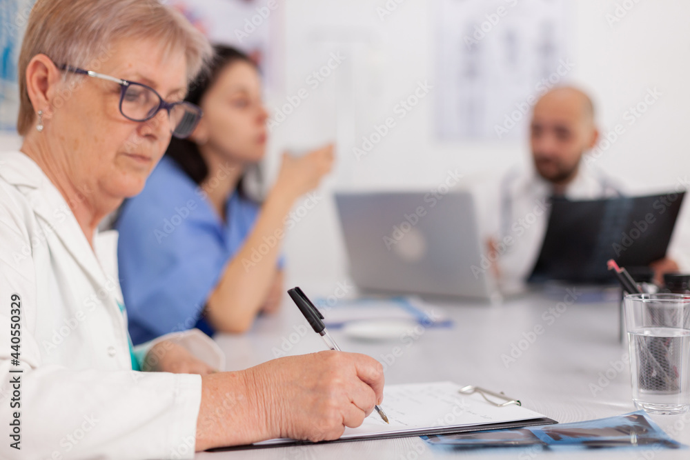 Physician senior woman in white coat analyzing medical examination prescribing pill medication writing sickness treatment on clipboard. Hospital teamwork sitting at desk in conference meeting room