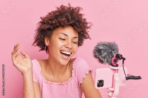 Positive pretty brunette Afro American female blogger focused at smartphone on tripod makes online streaming has own channel smiles gladfully poses against pink background. Vlogging concept. photo