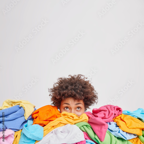 Young Afro American woman surrounded by different colorful clothes sorts out wardrobe focused above isolated over white background blank space for your advertising content. Nothing to wear concept