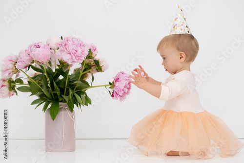 Adorale caucasian blonde baby girl wearing gala dress, birthday hat touching beautiful pion pink flower bouquet for holiday. Gift for kid, celerating idea, Greeting card photo