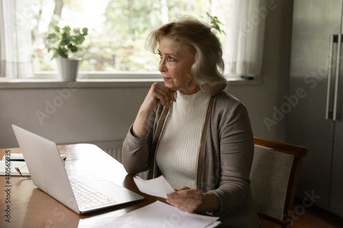 Pensive middle-aged 60s Caucasian woman work on laptop at home pay bills on online banking look in distance thinking. Thoughtful old female use computer busy with paperwork correspondence documents.