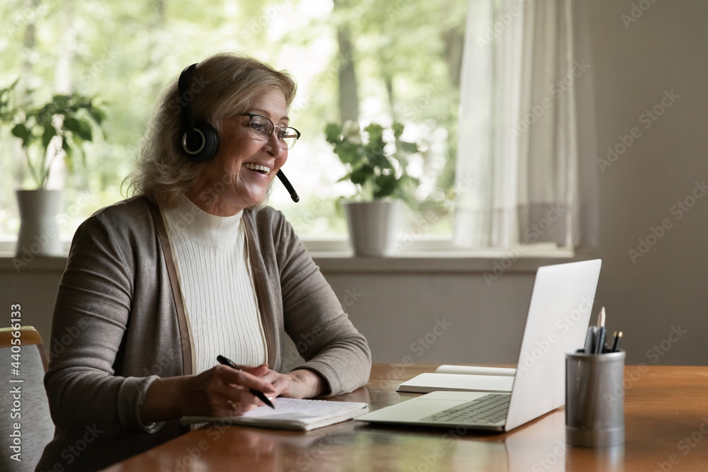 Smiling modern Caucasian old woman wear headset have fun use computer study online on gadget. Happy smart middle-aged female in earphones write note take distant course or training on laptop. Smiling modern Caucasian old woman wear headset have fun use computer study online on gadget. Happy smart middle-aged female in earphones write note take distant course or training on laptop.