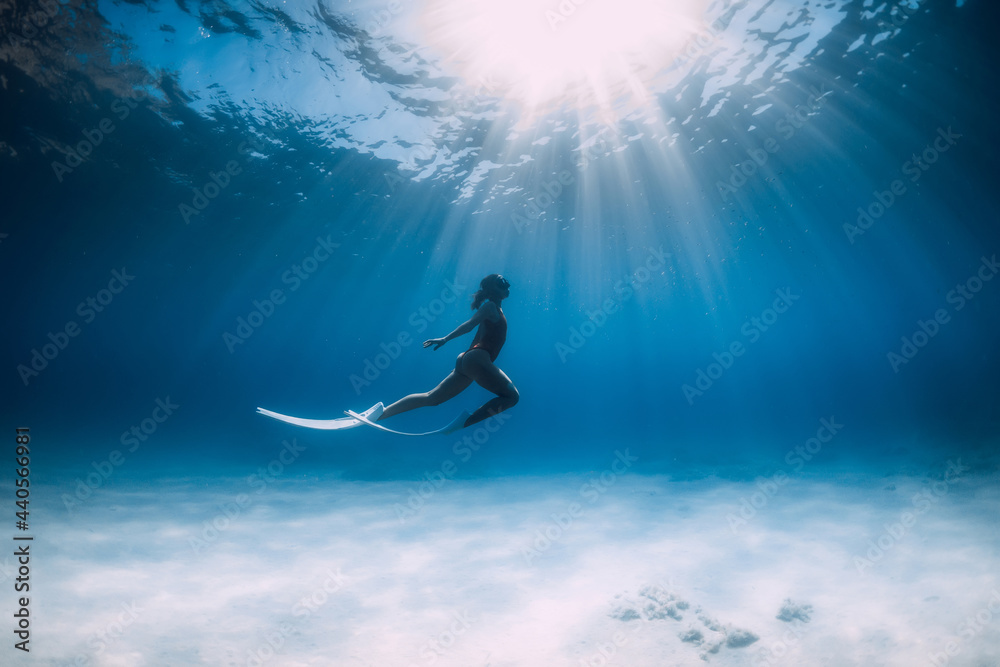 Freediver with white fins glides and posing underwater in sea with sunlight.