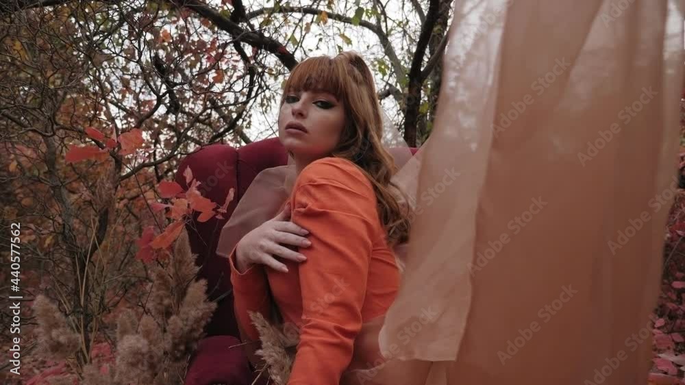 Young woman with long red hair posing in the autumn forest near a red soft chair Young woman with long red hair posing in the autumn forest near a red soft chair