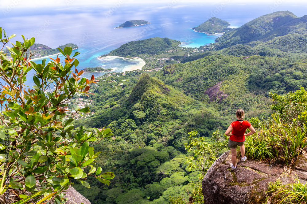 Young female traveller standing at the edge of the cliff at Morne Blanc View Point, overlooking Mahe Island coastline with lush tropical vegetation and crystal blue ocean, Seychelles. - obrazy, fototapety, plakaty 