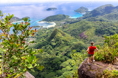 Young female traveller standing at the edge of the cliff at Morne Blanc View Point, overlooking Mahe Island coastline with lush tropical vegetation and crystal blue ocean, Seychelles. photo