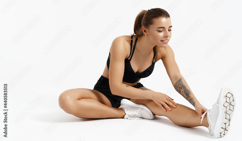 Image of young fitness woman, athlete sits on floor stretching leg before jogging. Runner prepare body for workout training session, smiling pleased, wearing summer activewear, white background