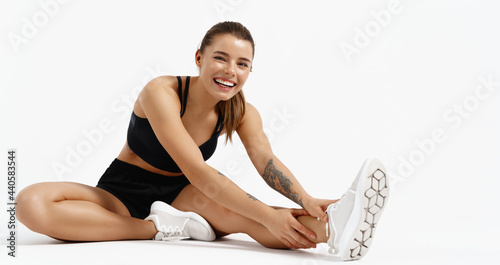 Smiling healthy fitness woman sitting on floor stretching leg, looking happy at camera. Gym girl athlete stretch and prepare body for workout, training session, white background photo