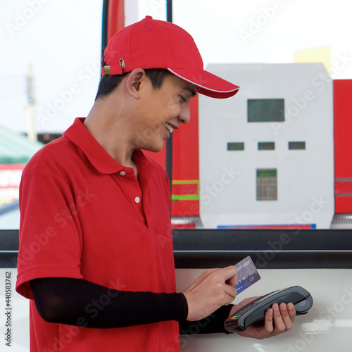 Gas station worker in red uniform stand smiling, swipe mockup credit card via payment terminal. Cash, price and volume readout display on petrol pump display screen. photo