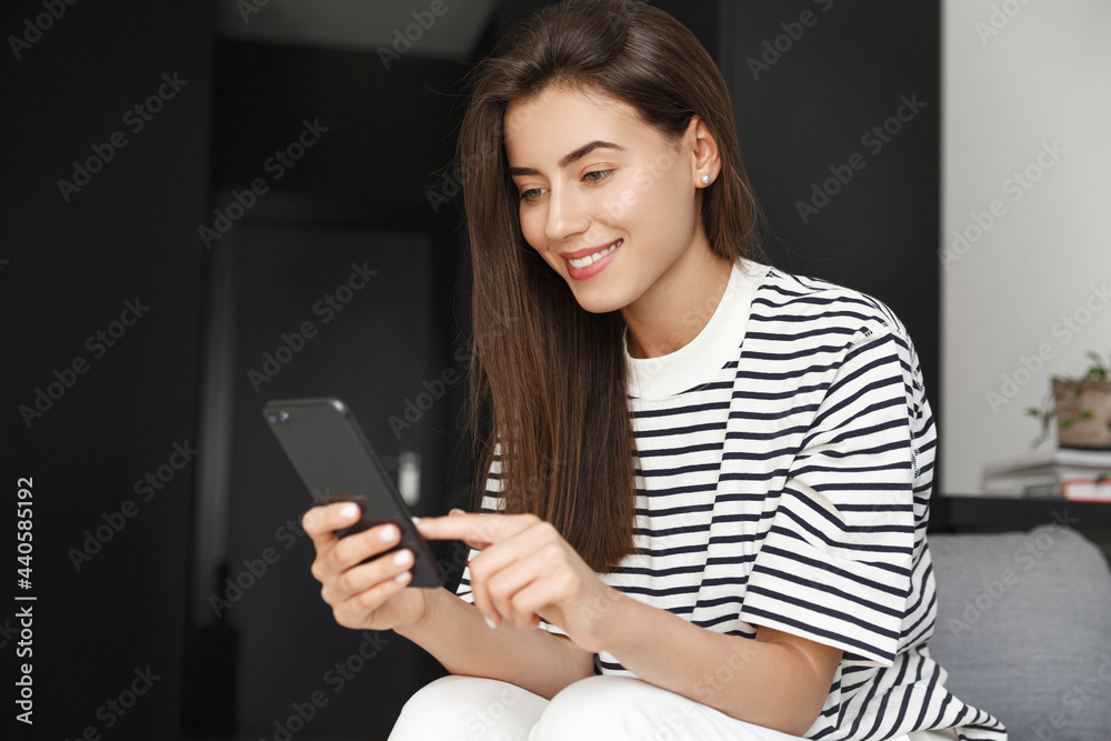 Foto Stock Beautiful girl using smartphone while sitting at home alone.  Girl relaxing in her house, networking, holding mobile phone and tap on  touch screen, choose smth or click link | Adobe