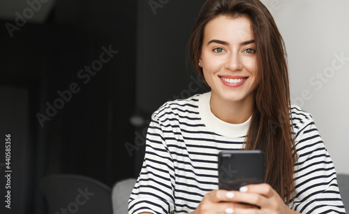Portrait of young happy woman using smartphone. Girl sitting at home and chatting, surf net on mobile phone, smiling at camera