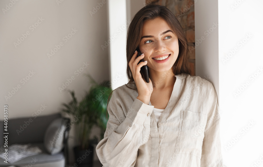 Young woman standing near office window, having phone call. Girl freelancer talking on smartphone, having conversation and smiling, answer customer, working remote Young woman standing near office window, having phone call. Girl freelancer talking on smartphone, having conversation and smiling, answer customer, working remote