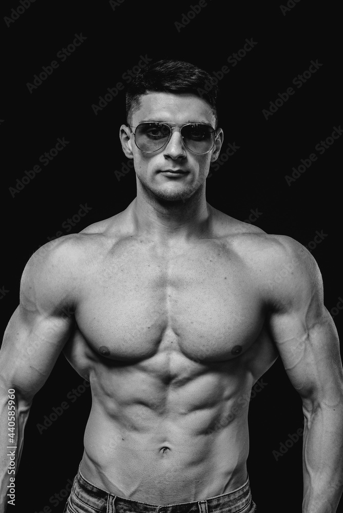 A young sexy athlete with perfect abs poses in the studio topless in jeans in a pair of protective glasses. Healthy lifestyle, training programs and nutrition for weight loss. Black and white.