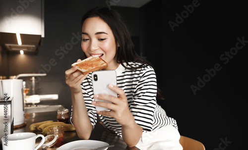 Young asian woman eating toast and watching video on smartphoone. Gil taking selfie while eat breakfast, blogger making content for social networks in kitchen photo