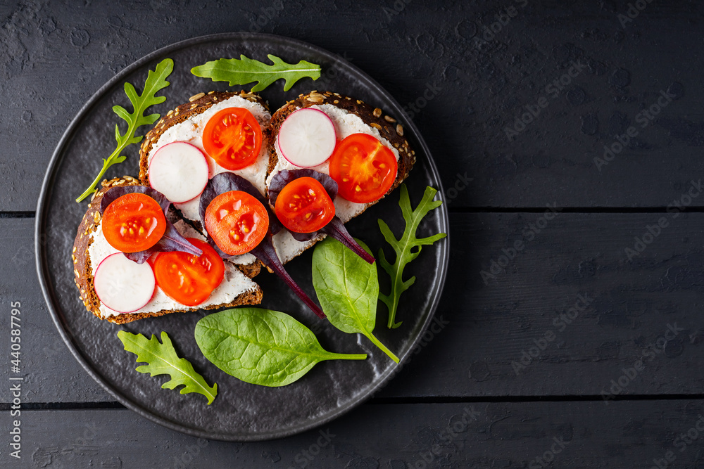 Toasts with cream cheese, cherry tomatoes and radish on black plate. Sandwiches with cream cheese and fresh herbs on a dark boards. Copy space. Flat lay