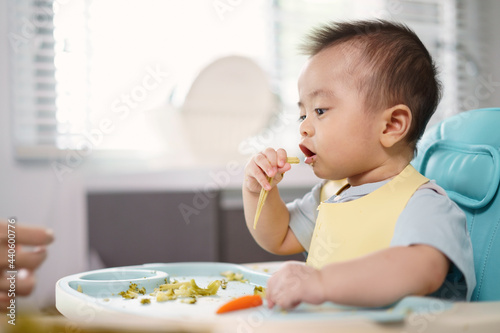 Asian little baby holding a baby corn. A son seat on a high chair in the kitchen at home. Finger food and healthy concept.