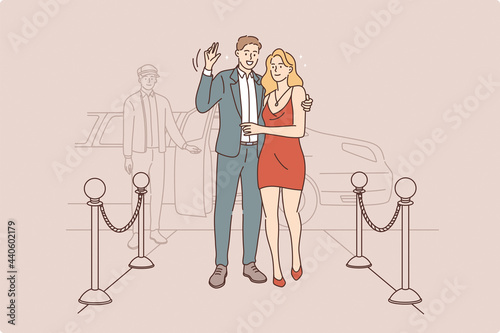 Famous couple waving to camera concept. Young smiling couple stars in elegant clothing standing in hotel entrance waving hands to camera with taxi awaiting behind 