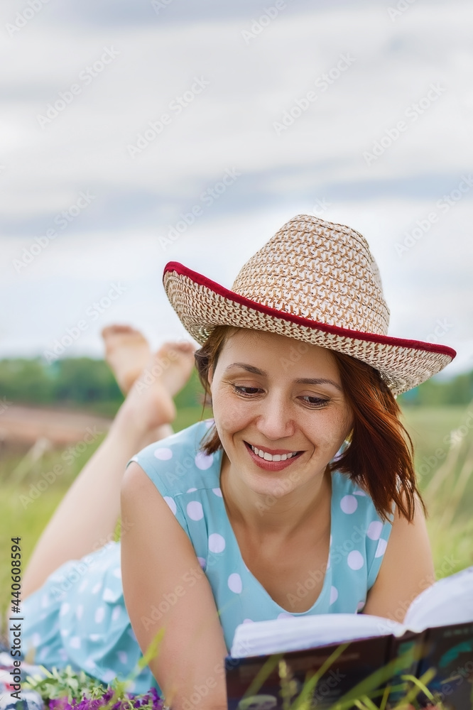 portrait of young woman in dress and summer hat lying in meadow and reading book in the warm summer day