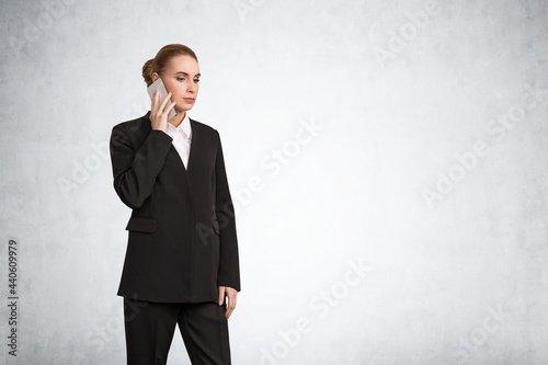 Attractive thoughtful businesswoman in formal black suit with white shirt is talking phone and pondering how to optimize business process to boost sales of the company. Concrete wall on background