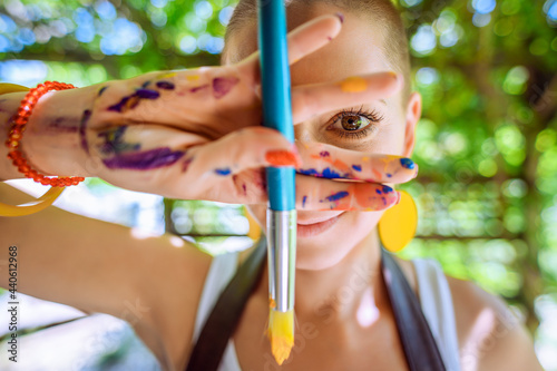 Playful portrait of a young gorgeous female painter artist, with hands covered in paint, looking and smiling at camera through her fingers. Creativity and individuality concept. photo