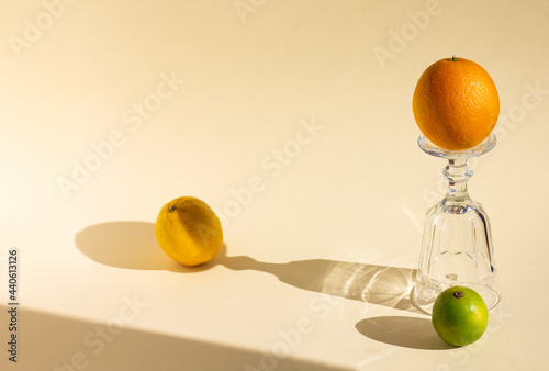 art creative composition of upside down empty goblet, orange, lemon and lime, crystal glass and fruits are under hard light on soft beige background, long glitter harsh shadows, high angle